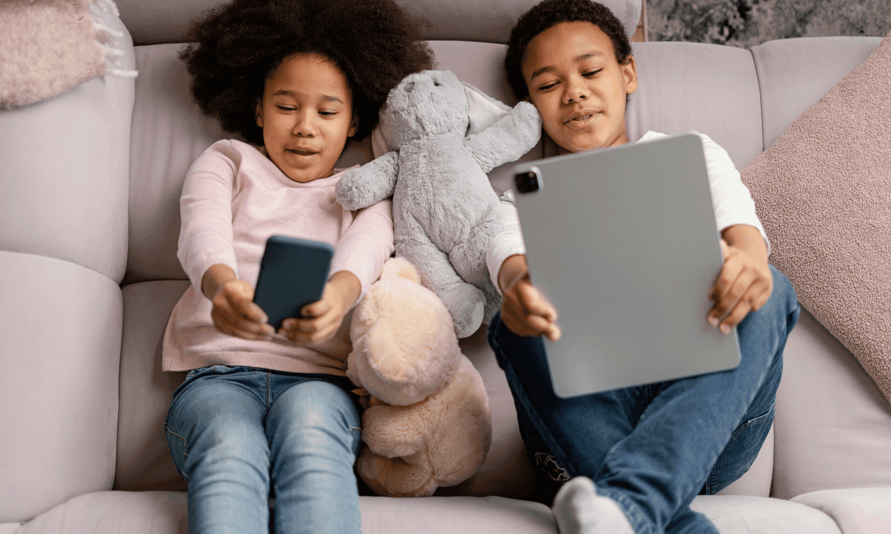 two kids laying on the sofa and holding a tablet and a mobile phone