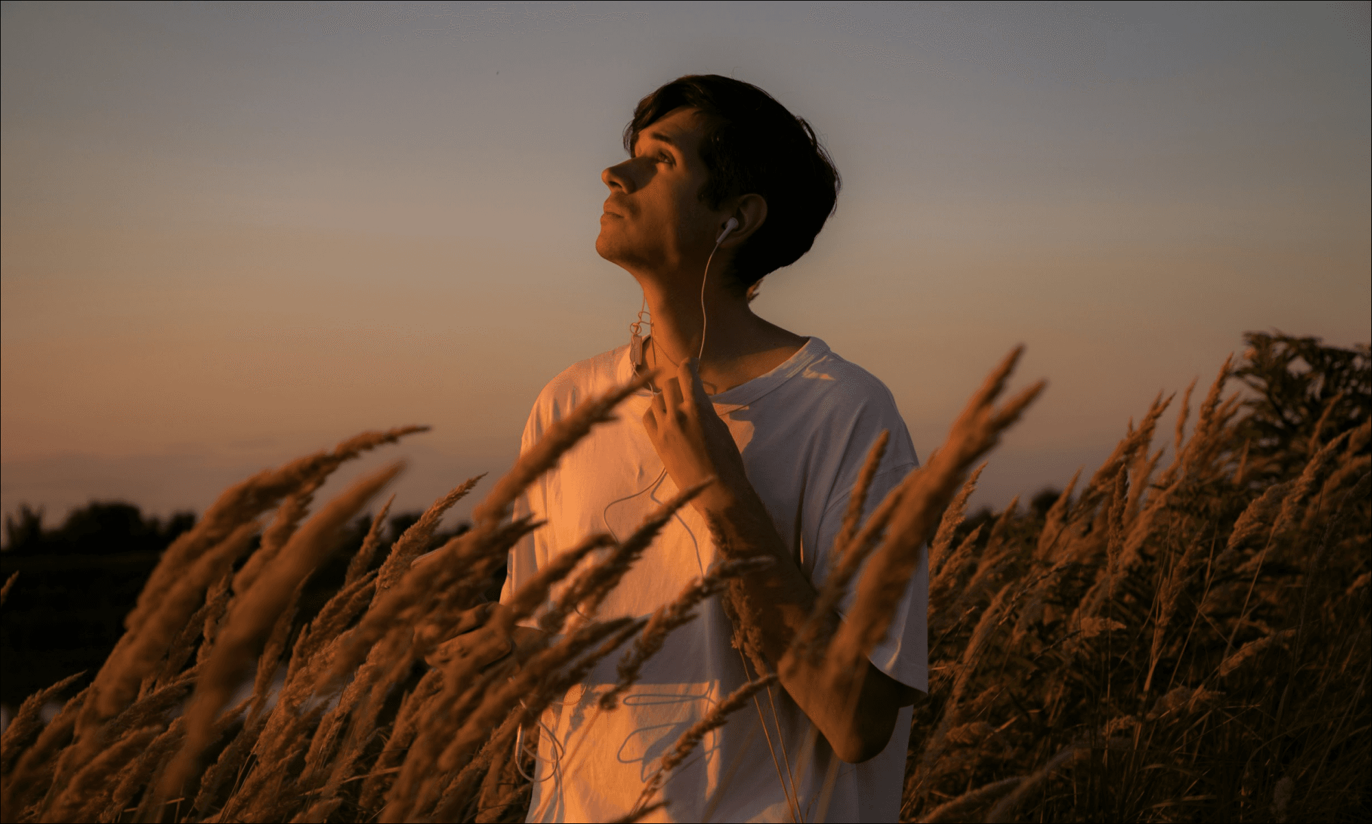 a man listening to music on his phone with wired headphones in a field at sunset